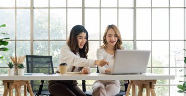 Masada dizüstü bilgisayarda çalışan iki Asyalı kadın. Arkaplan, yeşil doğanın manzarasına bakan camdan bir duvar.