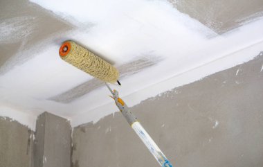 Yeni yenilenmiş bir odanın tavanına yeni bir kat beyaz boya sürmek için bir boya silindiri kullanılıyor. Duvarın pürüzsüz bir sonu var, son güncellemeyi gösteriyor.