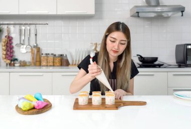 Önlüklü genç Asyalı bir kadın boru torbası kullanıyor, topkekin üzerindeki kremayı boruya yapıştırıyor, tahtadan bir tahtaya kek süslüyor, keklerin sunumuna renkli bir dokunuş ekliyor..