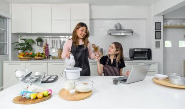 İki kişi ferah bir mutfakta birlikte çalışıyor. Biri mikser kullanırken diğeri dizüstü bilgisayarla etkileşime geçiyor. Pişirme projelerini hevesle planlıyorlar..