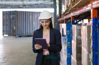 Takım elbiseli ve kasklı genç iş kadını tablet bilgisayardaki envanter kayıtlarını inceliyor. Yoğun bir depolama alanında istiflenmiş kutuların arasında dur. Konteynır doldurma ve kargo koruma servisinde çalışıyorum..