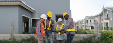 Dört inşaat işçisi planlarını tablet bilgisayarda tartışmak ve inşaat alanının dışında durmak için toplandı. Kask ve güvenlik yeleği giyiyorlar, çevre yapılar arasında görevlerine odaklanıyorlar.