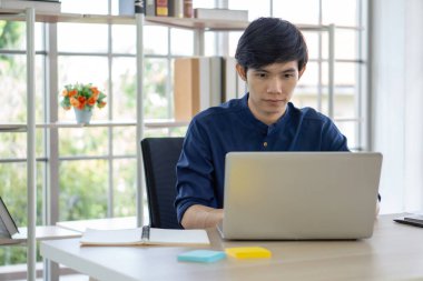 Bilgisayarına odaklanmış genç bir adam notlar ve renkli notlarla dolu çağdaş bir masada oturmuş, büyük pencereden akan doğal gün ışığının tadını çıkarıyordu..