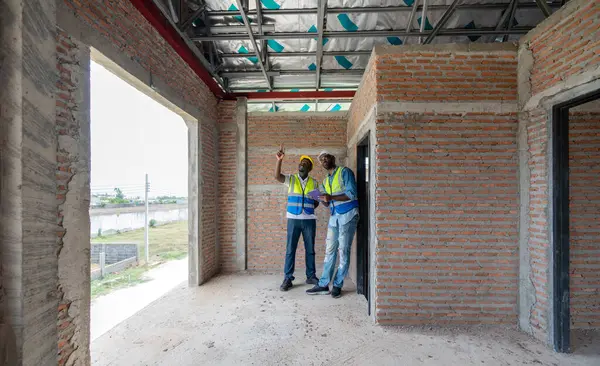 Güvenlik yeleği giymiş iki inşaat işçisi ve tablet bilgisayarı tutarken inşaat çalışmalarını denetliyor. Gündüz vakti gelişmekte olan bir yerleşim bölgesindeki inşaat alanında atmosfer