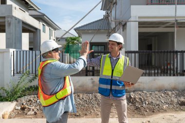 İki inşaat işçisi bir inşaat sahasında dostça bir değiş tokuş yapıyor, açık havada plan ve ilerlemeyi tartışıyorlar. Atmosfer işbirlikçi ve üretken..