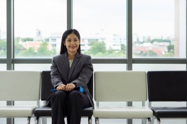 Gri takım elbiseli genç Asyalı iş kadını pencerenin yanındaki bankta oturuyor, kucağında mavi bir dosya var..