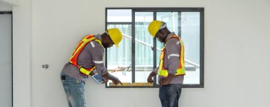 Güvenlik teçhizatlı iki inşaat işçisi binanın içindeki bir kat aletini kullanarak bir pencere kurulumunu dikkatlice değerlendiriyor. Ayar parlak ve profesyonelce, gayret ve güvenliği yansıtıyor..