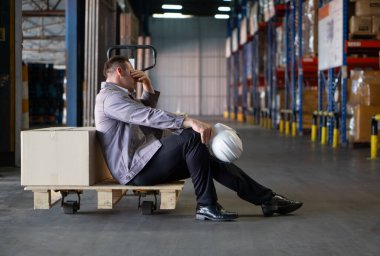 Bir depo işçisi karton kutularla çevrili ahşap bir paletin üzerinde dinleniyor. Yorgun görünüyor, elinde sert bir şapkayla, yoğun bir iş gününü yansıtıyor..