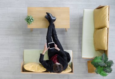 Takım elbiseli genç işadamı kanepeye oturur, ayaklarını masaya koyar ve ilginç yatırım bilgileri için tablet bilgisayar kullanır. Üst Görünüm