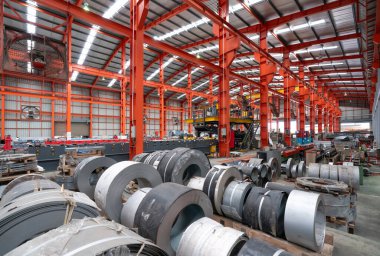 Kocaman bir depo, kafasının üzerinden geçen parıldayan çelik kirişlerden oluşan bir labirentle dolup taşarak metalik bir tepe örtüsü oluşturuyor. Yerde çelik levhalar ve metal levhalar var..