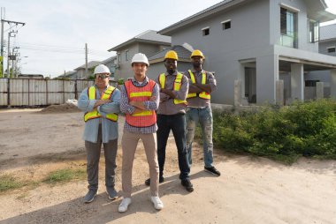 İnşaat işçisi grubu konut projelerinin şantiyesinde kolları bağlı duruyor. Kask ve çelik yelek giyiyorlar.