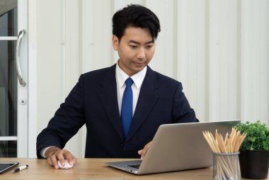 Takım elbiseli profesyonel bir adam, çağdaş bir ofiste ahşap bir masada oturup dizüstü bilgisayarına odaklanır..