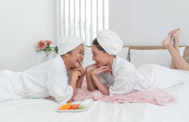 Gülümseyen ve beyaz bornoz giyen iki Asyalı kadın, çiçeklerle süslenmiş parlak bir odada rahat bir yatakta dinleniyorlar. Bir sağlık kaçamağı sırasında birbirlerine eşlik etmekten hoşlanırlar..