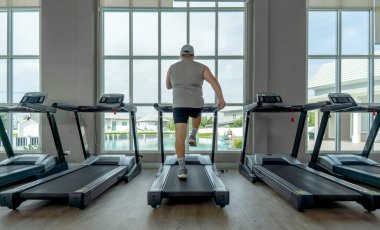 Spor salonunda koşu bandında yürüyen bir adam. Pencerenin önünde dur, dışarıdaki havuzun tadını çıkar. Parlak güneş ışığı spor salonunu dolduruyor ve egzersiz için davetkar bir atmosfer yaratıyor..