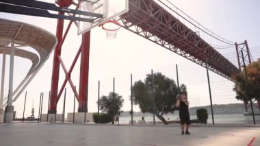 Güneşli bir günde bir adam, arka planda bir nehir üzerinde köprüsü olan bir basket topunu potaya atar ve sonra çerçeveden çıkıp gider..