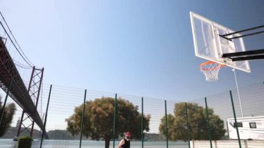 Güneşli bir günde bir adam, arka planda bir nehir üzerinde köprüsü olan bir basket topunu potaya atar ve sonra çerçeveden çıkıp gider..