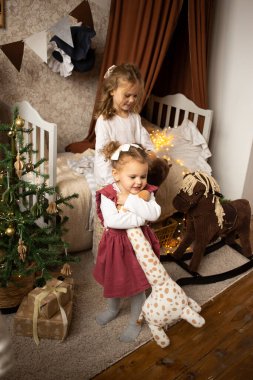 İki kız kardeş çocuk odasında, yatağın yanında oyuncak hayvanlarla oynuyorlar.