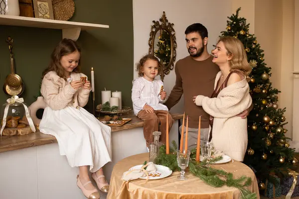 両親と2人の姉妹の子供たちがキッチンでクッキーを持っています 背景にクリスマスツリー ロイヤリティフリーのストック画像