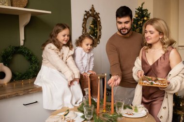 Anne, baba ve evde kurabiyeli iki kız kardeş. Arka planda Noel ağacı.