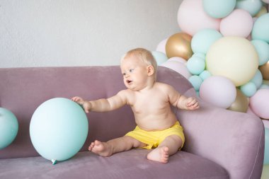 Evdeki koltukta şort giymiş şirin bir oğlan çocuğu renkli bir balonun arka planında oturuyor.