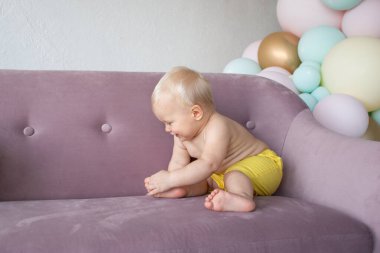 Şirin, küçük, gülümseyen bir çocuk. Evdeki koltukta, renkli balonların arka planında oturuyor.