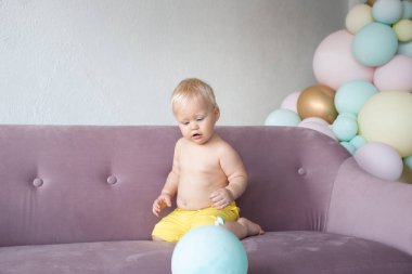 Şirin tombul bir oğlan. Evdeki koltukta şort giymiş renkli balonların arka planında oturuyor.