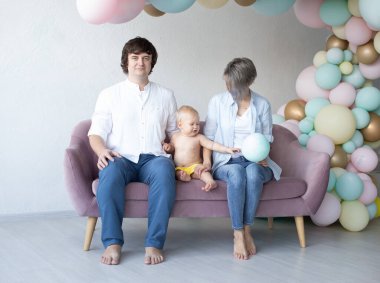 Anne, baba ve oğulları ilk doğum gününde renkli balonların arka planında kanepede oturuyorlar.