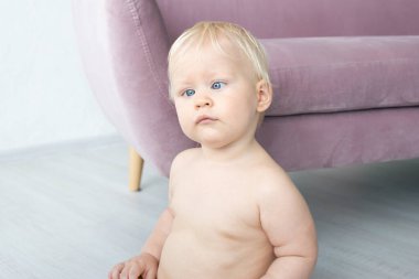 Strabismus gözlü, evde şort giymiş, yerde oturan tatlı bir erkek bebek.