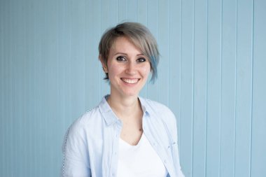 Mavi ahşap bir duvarın yanında duran ve evinde gülümseyen kısa saçlı şık bir kadın.