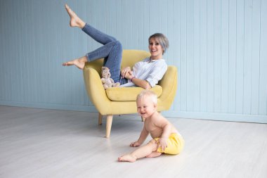 Mutlu genç bir anne, evdeki koltukta oturan oğluyla oynuyor. Mavi tahta bir duvarın yanında, mutlu bir aile.