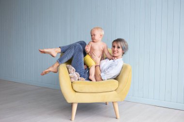 Mutlu genç bir anne Strabismus gözlü oğluyla oynuyor. Evdeki koltukta oturuyor. Mavi ahşap duvarın yanında, mutlu bir aile.