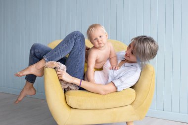 Mutlu genç bir anne Strabismus gözlü oğluyla oynuyor. Evdeki koltukta oturuyor. Mavi ahşap duvarın yanında, mutlu bir aile.