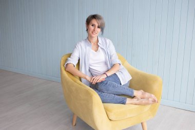 Evdeki koltukta oturan kısa saçlı kadın. Gömlekli ve kot pantolonlu rahat bir kadın portresi koltukta dinleniyor. Gülümseyen ve rahatlatan güzel bir kızın portresi