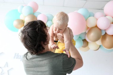 Mutlu genç baba, yeni yürümeye başlayan küçük erkek çocuğuyla oynuyor. Mutlu modern aile renkli balonların yanında