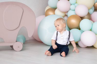 İlk doğum gününde evde takım elbiseli, barfiks pantolonlu ve renkli balonların arka planında bir gömlek ve oyuncak bir fil olan sevimli bir çocuk. Aile ve çocukluk kavramı, tatil