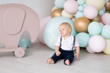 İlk doğum gününde evde takım elbiseli, barfiks pantolonlu ve renkli balonların arka planında bir gömlek ve oyuncak bir fil olan küçük bir çocuk. Aile ve çocukluk kavramı, tatil