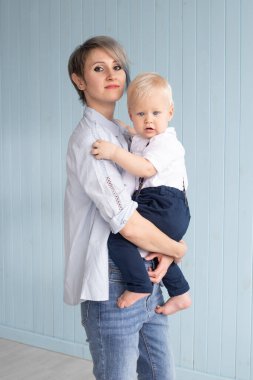 Elinde oğlu olan genç bir anne, kısa saçlı bir kadın mavi ahşap bir duvarın yanında duruyor ve evde gülümsüyor, mutlu bir aile.