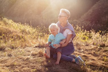 Oğlan ve annesi yazın gün batımında güneş ışınlarıyla çimenlerde otururlar.