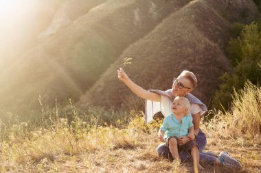 Anne ve küçük oğlu yazın doğada oynuyorlar çimenlerde oturuyorlar gün batımında günbatımında yeşil tepelerin yakınında