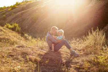 Genç mutlu anne yazın doğada oğlunu kucaklıyor çimlerin üzerinde oturuyor gün batımında yeşil tepelerin yakınında güneş ışınlarıyla.