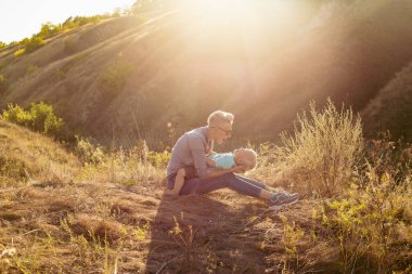 Genç mutlu anne yazın doğada oğlunu kucaklıyor çimlerin üzerinde oturuyor gün batımında yeşil tepelerin yakınında güneş ışınlarıyla.