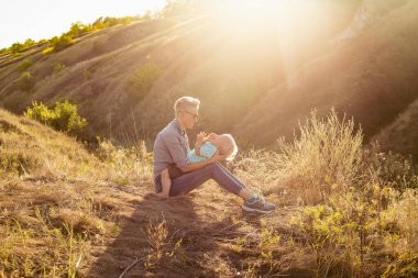Genç anne oğluyla birlikte yazın doğada oynarken çimlerin üzerinde güneş ışınlarıyla yeşil tepelerin yakınında oturuyor.