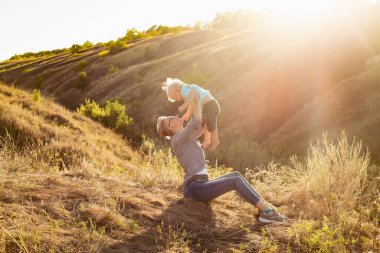 Anne, yazları doğada oğlunu kaldırıyor çimlerin üzerinde oturuyor gün batımında yeşil tepelerin yakınında güneş ışınlarıyla.