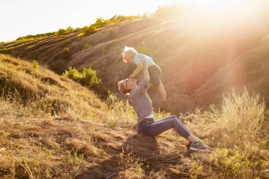 Anne gülümsüyor ve doğada küçük oğlunu kaldırıyor. Yazın çimlerin üzerinde oturuyor. Gün batımında yeşil tepelerin yakınında güneş ışınlarıyla oturuyor.