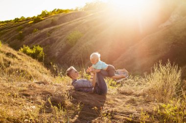 Anne gülümsüyor ve oğlunu doğada doğada kaldırıyor. Yeşil tepelerin yakınında güneş ışınlarıyla çimlerin üzerinde oturuyor.