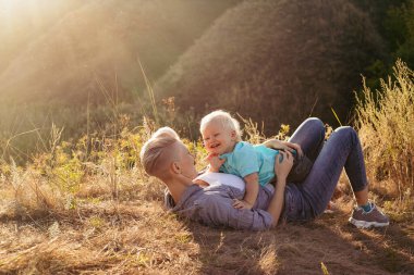 Anne gülümsüyor ve oğlunu kucaklıyor yazın çimlerin üzerinde oturuyor gün batımında yeşil tepelerin yakınında güneş ışınlarıyla