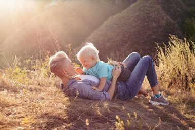 Anne gülümsüyor ve oğlunu kucaklıyor yazın çimlerin üzerinde oturuyor gün batımında yeşil tepelerin yakınında güneş ışınlarıyla