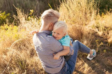Genç mutlu anne yazın doğada oğlunu kucaklıyor çimlerin üzerinde oturuyor gün batımında yeşil tepelerin yakınında güneş ışınlarıyla.