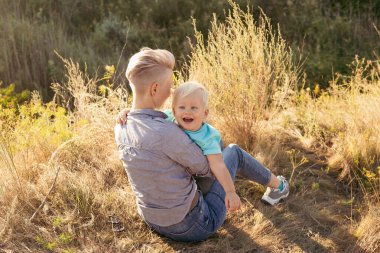 Anne gülüyor ve oğlunu kucaklıyor yazın çimlerin üzerinde oturuyor gün batımında yeşil tepelerin yakınında güneş ışınlarıyla