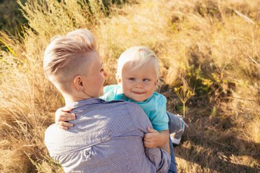 Anne, yazın doğada oğlunu kucaklıyor çimlerin üzerinde oturuyor gün batımında yeşil tepelerin yakınında güneş ışınlarıyla.
